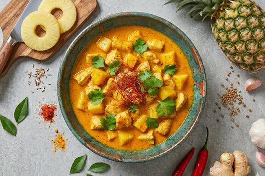 Ein Ananas Curry mit Poulet und cremiger Kokossauce steht mit frischer Ananas, Koriander und Gewürzen auf hellem Untergrund