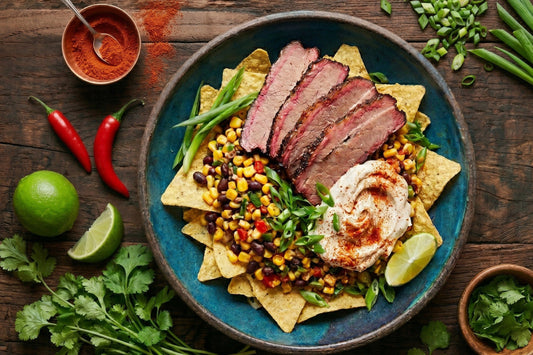 Ein Teller mit Brisket Tacos auf Nachos, dazu Mais-Bohnen-Salsa, Limetten und Creme – ein BBQ-Rezept mit südamerikanischem Charakter