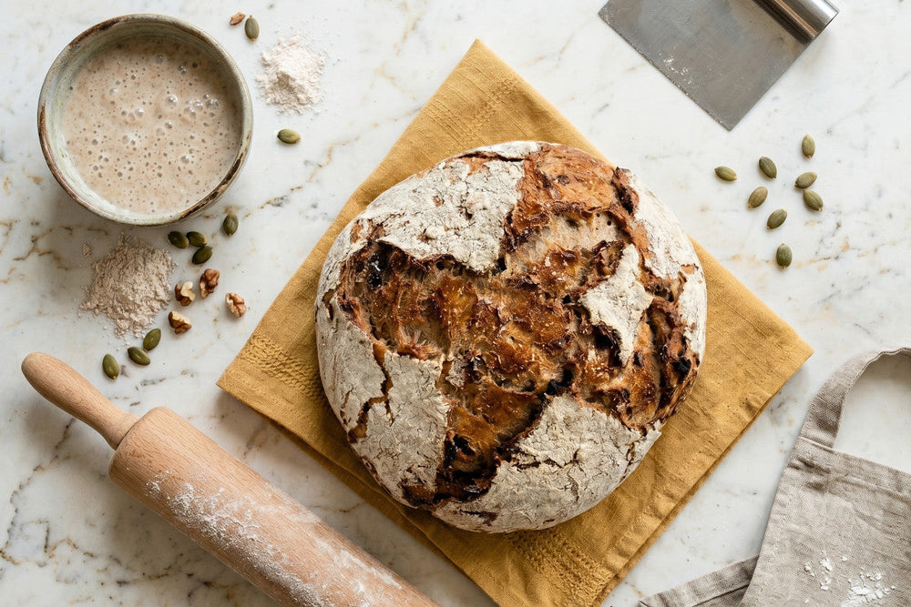 Ein frisch gebackenes Roggenvollkorn-Sauerteigbrot liegt mit Sauerteig, Kernen, Mehl und Wallholz auf hellem Untergrund