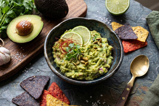 Ein cremiger Avocado Guacamole Dip steht mit Limette, Kürbiskernen, Koriander, Avocado und Tortilla-Chips auf dunklem Stein