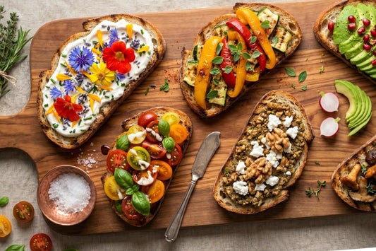 Mehrere Bruschetta-Varianten liegen auf einem Holzbrett, belegt mit Tomaten, Grillgemüse, Avocado, Kräutern und Blüten