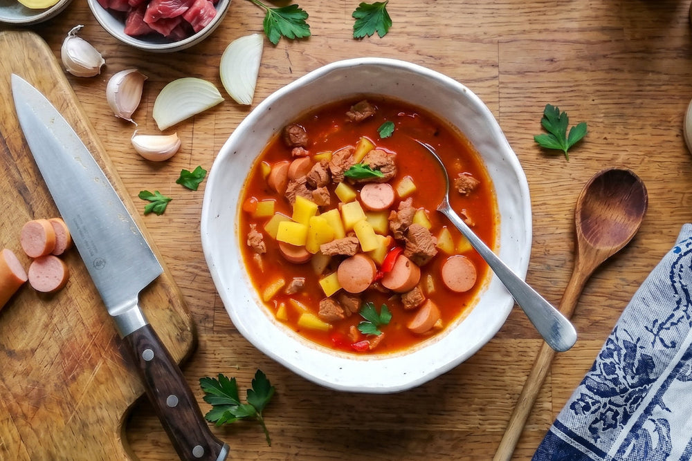 Eine herzhafte Gulasch Suppe mit Fleisch, Kartoffeln, Wurst, Peperoni und Kräutern steht auf einem Holztisch