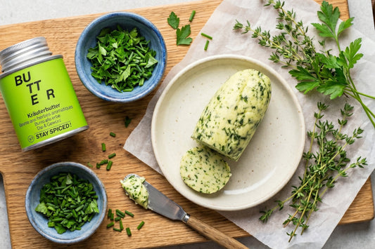 Eine Rolle Kräuterbutter liegt auf einem Teller, umgeben von frischen Kräutern, Messer und Gewürzdose