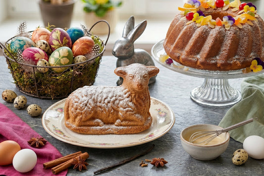 Ein Osterlamm Kuchen steht mit Puderzucker, bunten Ostereiern, Gewürzen und einem Gugelhupf auf einer festlichen Tafel