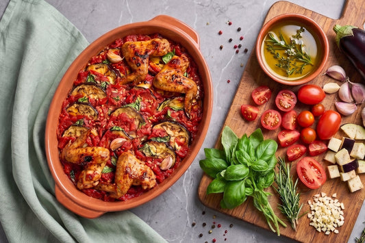 Ein Poulet-Auberginen Auflauf mit Tomaten, Basilikum und Kräuteröl liegt in einer Ofenform neben frischen Zutaten