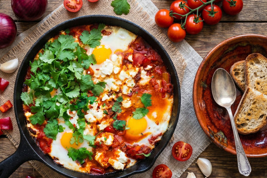 Eine Shakshuka mit Eiern, Tomatensauce, Feta und Koriander steht in einer Pfanne neben Tomaten, Brot und Gewürzen