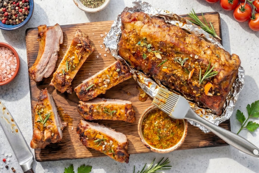 Glasierte Spareribs liegen mit Kräutern, Marinade und Pinsel auf einem Holzbrett und zeigen ein Rezept für saftige Rippchen