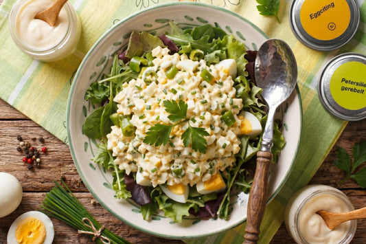 Cremiger Eiersalat mit Cornichons und Schnittlauch liegt auf Blattsalat, daneben Ei, Mayonnaise und Gewürzdosen